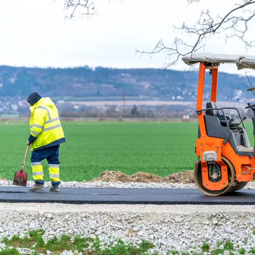 Ein Straßenarbeiter, der eine rote Schaufel in der Hand hält, blickt auf den frisch gepressten Asphalt einer neu gebauten Straße. Im Hintergrund erstreckt sich ein grünes Ackerfeld bis zu einem bewaldeten Hügel in der Ferne.